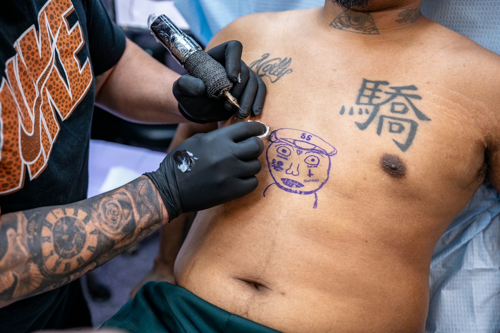 Tattoo artist inks a face onto a man's chest.